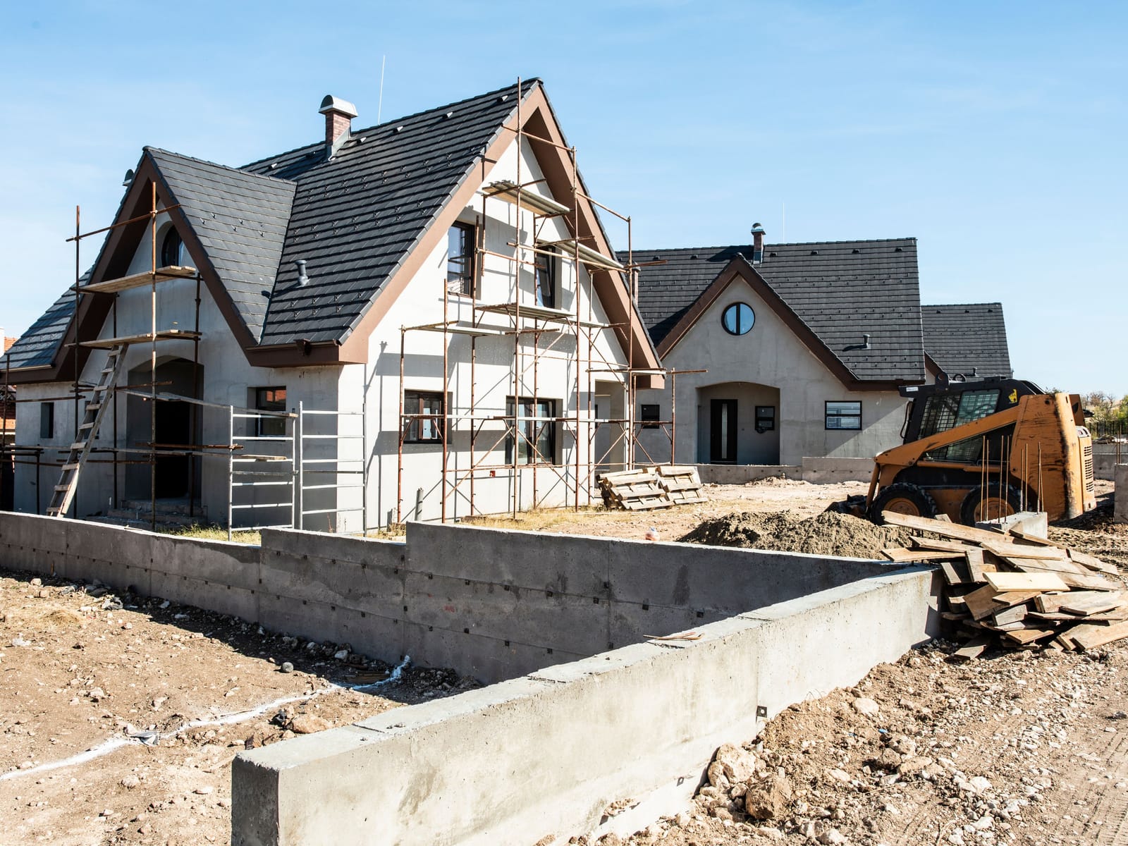 New homes under construction with scaffolding, a poured foundation wall, and a skid steer on site