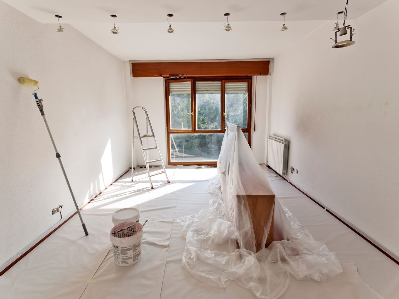 Room prepared for painting with drop sheets over the floor, furniture covered in plastic, a ladder, and a roller on an extension pole